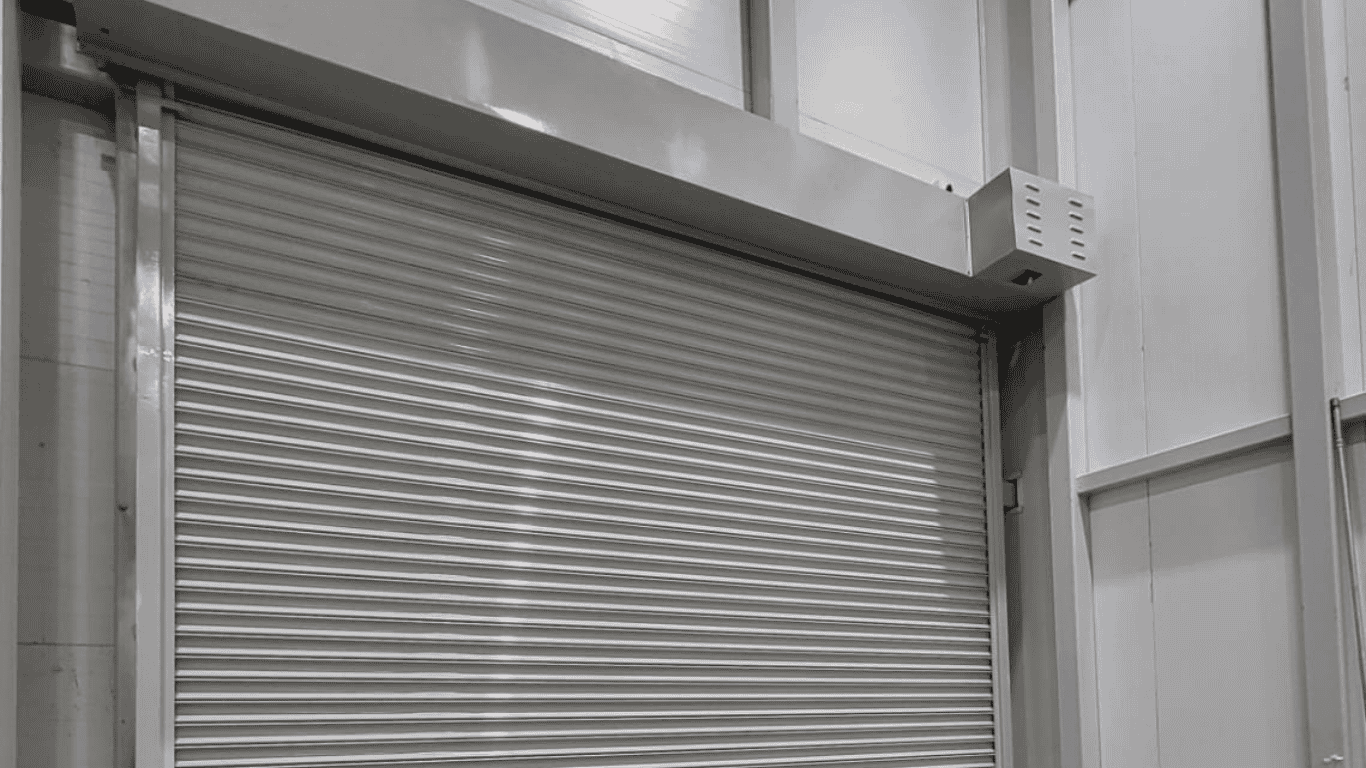 A closed industrial motorized rolling shutter door with horizontal panels in a large, modern building. The walls and ceiling are smooth and feature a light, neutral color. A rectangular vent box is mounted above the door on the right.